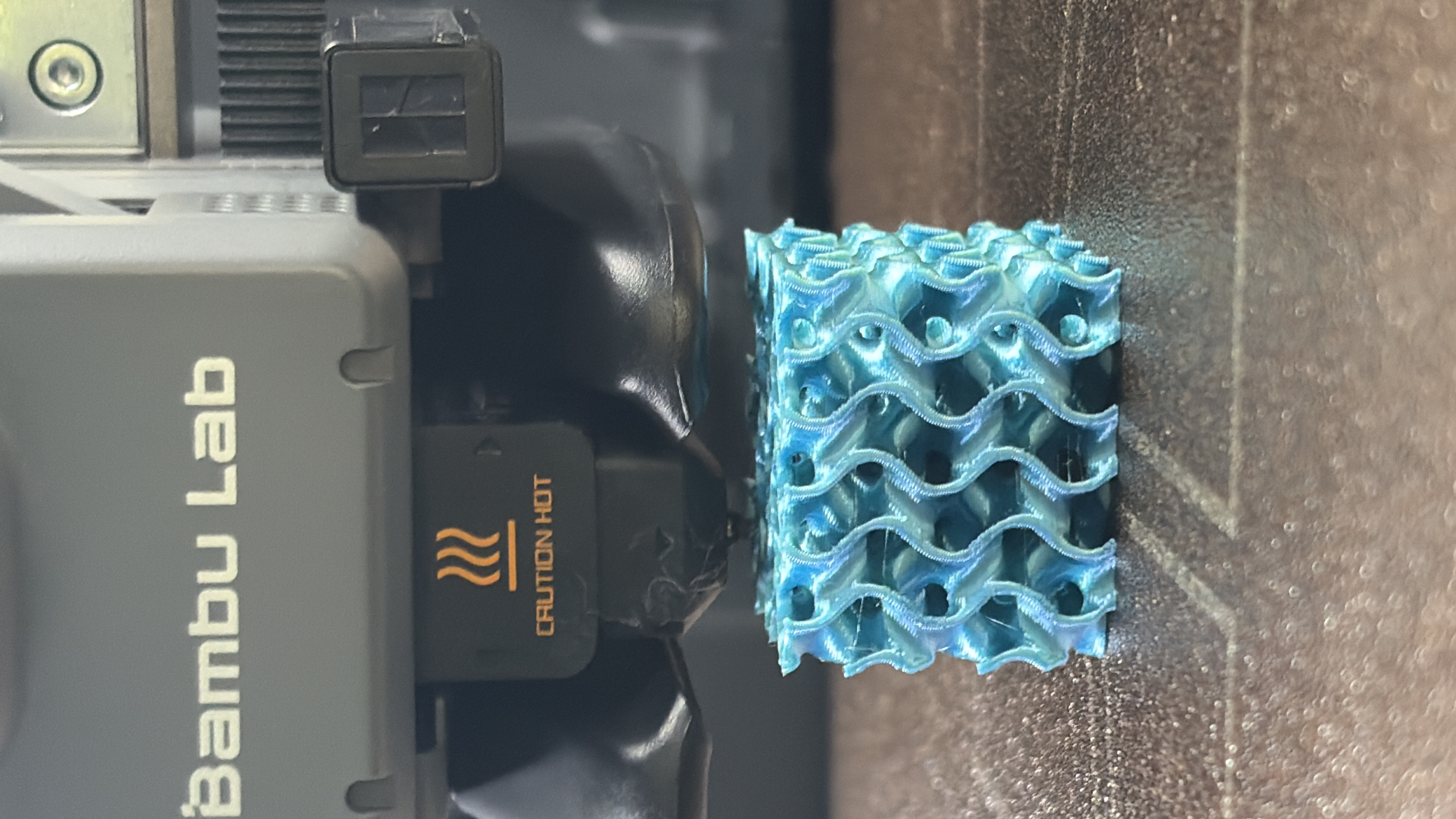 Freshly printed gyroid lattice cube in teal PLA, still under the Bambu Lab nozzle